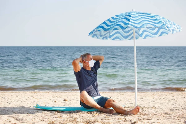 Fifty-year-old man with protective mask stands at the beach under an umbrella near the sea