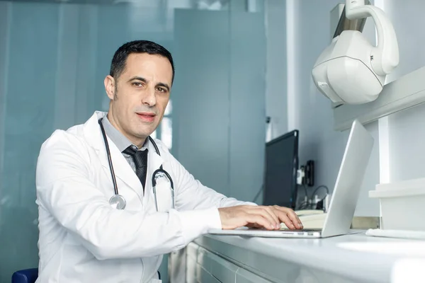 Dark-haired doctor with black hair in a white coat, working smiling at the computer in his surgery