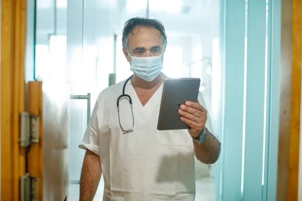 Doctor in a white coat and surgical mask looks at a tablet as he walks in a hospital ward