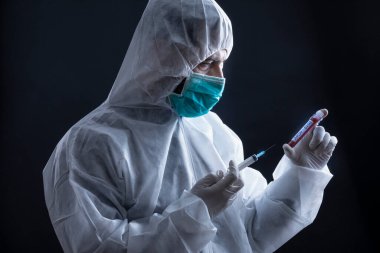 Koruyucu giysili ve maskeli doktor koronavirüs kan testi tüpü ve şırınga tutuyor. Siyah arka plan.