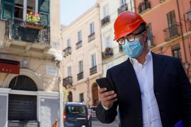Mühendis, cep telefonunu kullanırken koruyucu kask ve cerrahi maske takan bir inşaat alanının yanında duruyor.