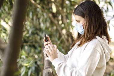 Yüz maskesi ve beyaz kazaklı kız cep telefonunu kullanırken parkta öylece duruyor.