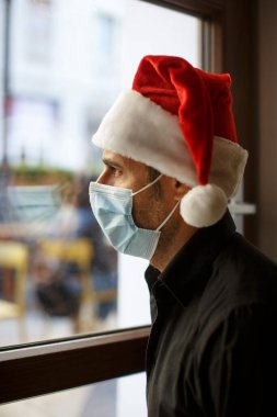 Noel Baba şapkalı ve koruyucu maskeli çocuk evin penceresinden üzgün görünüyor.