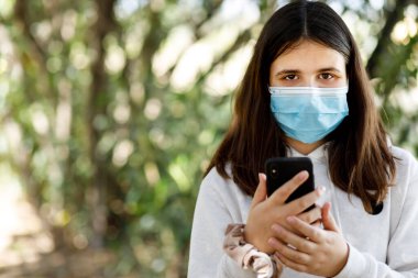 Yüz maskeli ve beyaz kazaklı kız cep telefonuyla sabit duruyor.