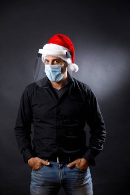 Siyah gömlekli, cerrahi maskeli, pleksiglas koruyucu kalkanlı ve Noel Baba şapkalı adam ciddi görünüyor, siyah arka planda izole edilmiş.