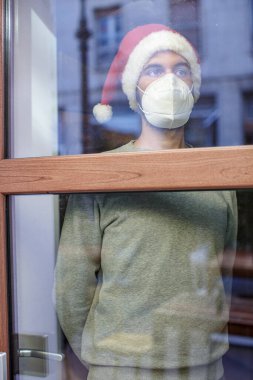 Noel zamanı pencereye bakan maskeli genç bir adam.