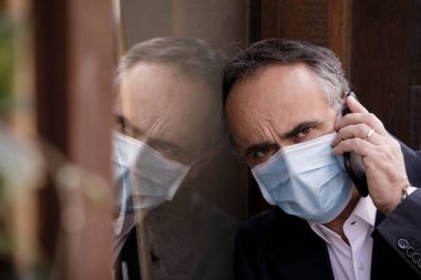 Ceketli ve maskeli bir adam, bardağa yansıyor, telefonda endişeli endişeli konuşuyor.
