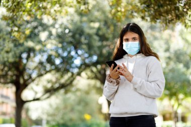 Yüz maskesi ve beyaz kazaklı kız cep telefonunu kullanırken parkta öylece duruyor.