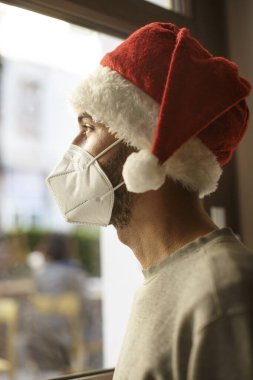 Noel Baba şapkalı ve koruyucu maskeli çocuk evin penceresinden üzgün görünüyor.