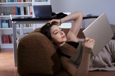 Güzel esmer kız tabletinden film izliyor. Evinin kanepesine rahatça sığıyor.