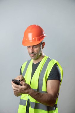 Turuncu koruyucu miğferi ve sarı yeleği olan işçi gri arka planda izole edilmiş cep telefonundan mesaj yolluyor.