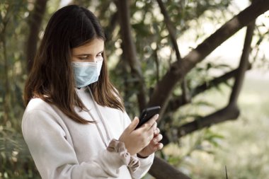 Yüz maskesi ve beyaz kazaklı kız cep telefonunu kullanırken parkta öylece duruyor.