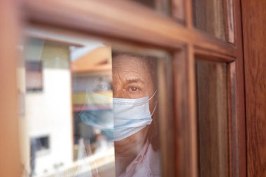 Yüzünde tıbbi maske olan yaşlı bir kadın pencereden dışarı bakıyor.