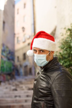 Noel Baba şapkalı, deri ceketli ve cerrahi maskeli adam şehir ortamında üzgün görünüyor.