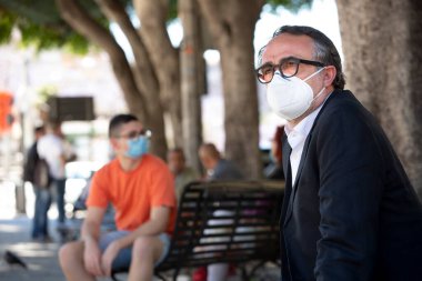 Şehir parkındaki banklarda oturan koruyucu maskeli genç ve yetişkin adamlar.