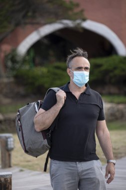 Yazlık kıyafetli ve sırt çantalı koyu renk saçlı bir adam koruyucu maskesiyle sahil kasabasında yürüyor.