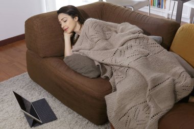 Koyu renk saçlı kız, battaniyesiyle kanepede yatarken halıya yerleştirilmiş tabletine bakar.