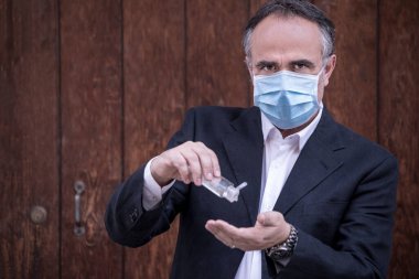 Takım elbiseli, koruyucu maskeli bir adam ahşap arka planda el dezenfektanı kullanıyor.