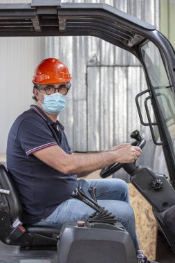 Koruyucu miğferi ve cerrahi maskesi olan bir işçi şirketinin içinde bir forklift kullanıyor.