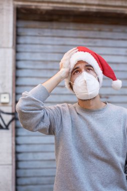 Noel Baba şapkası ve sağlık maskesi takan üzgün genç bir adam.