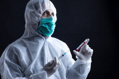 Koruyucu giysili ve maskeli doktor koronavirüs kan testi tüpü ve şırınga tutuyor. Siyah arka plan.