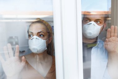 Maskeli kadın pencerenin arkasından eve bakıyor.