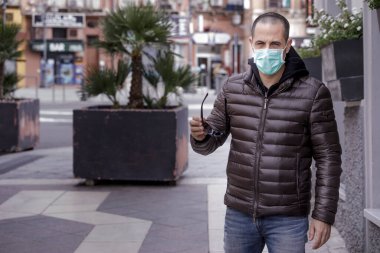 Koyu renk yelekli adam şehirde dolaşırken koruyucu bir cerrahi maske takıyor.
