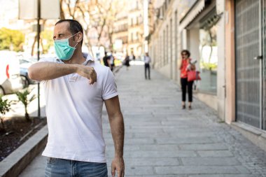 Siyah saçlı adam beyaz polo ve yüz maskeli hapşırıkları ile dirseğini kaldırımda sabit bir korumaya getiriyor.