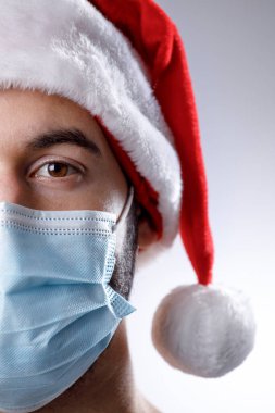 Ciddi bir çocuğun portresi, Noel Baba kılığında, cerrahi maskeli, beyaz arka planda izole edilmiş.