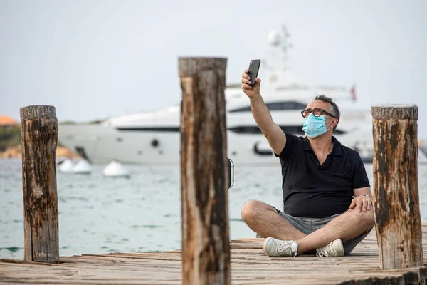 顔のマスクをした男 黒いシャツは足を組んで座っていて 海港の埠頭でスマートフォンで自撮りしている — ストック写真