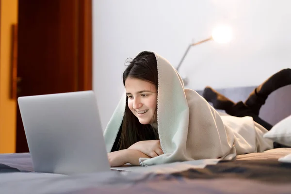 一个黑头发的小女孩在电脑上笑着躺在床上 头上蒙着毯子 — 图库照片