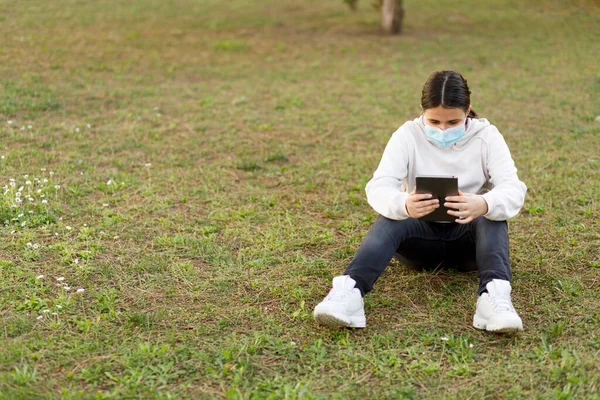 Yüz Maskeli Genç Bir Kız Parkta Tabletini Kullanıyor — Stok fotoğraf