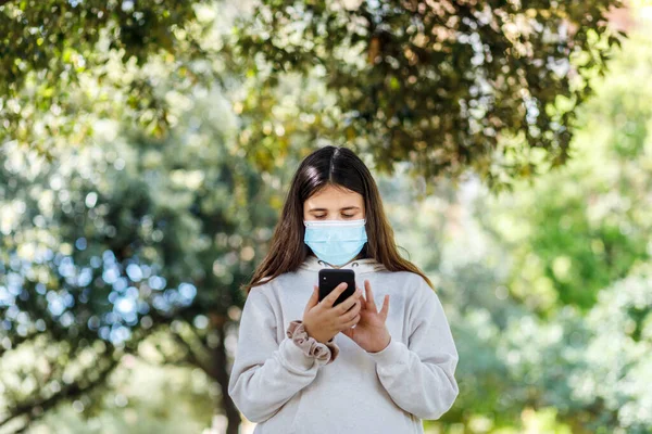 Yüz Maskesi Beyaz Kazaklı Kız Cep Telefonunu Kullanırken Parkta Öylece — Stok fotoğraf
