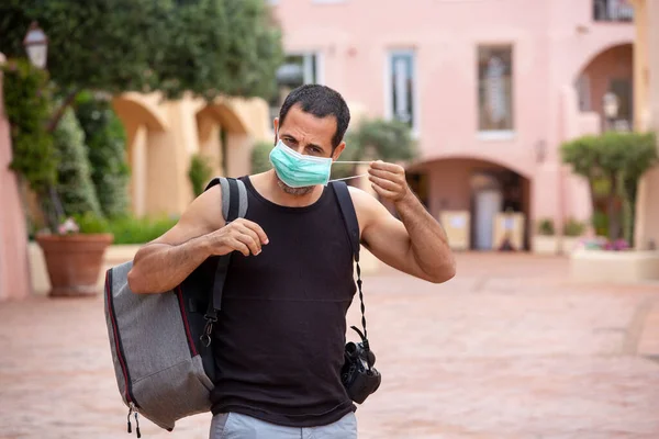 夏の服装の黒髪の男とバックパックは保護顔のマスクを持つリゾートタウンを歩き回ります — ストック写真