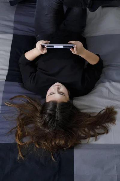 小女孩躺在地上 一头乌黑的头发绑在背后 一件黑色的衬衫正在看她平板电脑上的一部电影 — 图库照片