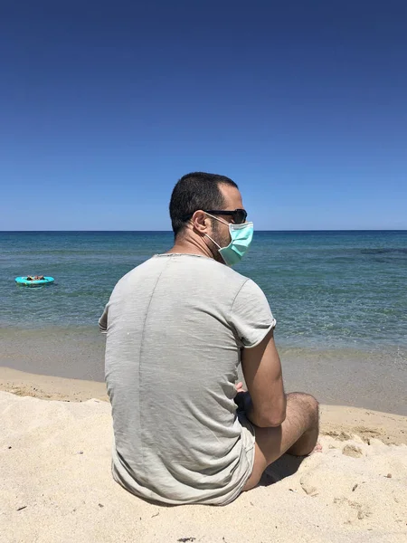 眼鏡とTシャツを着た黒髪の男の子が海岸の海の前に座っている防護外科用マスクを着て — ストック写真