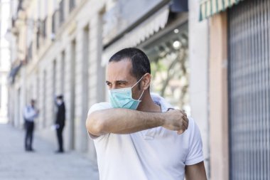 Siyah saçlı adam beyaz polo ve yüz maskeli hapşırıkları ile dirseğini kaldırımda sabit bir korumaya getiriyor.