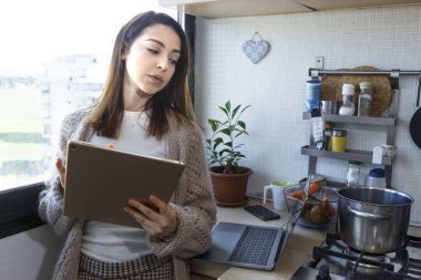 Mutfakta rahat giyimli bir kız yemek pişirirken tabletine bakıyor.