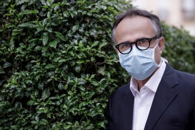 Ceketli, koruyucu maskeli, orta yaşlı bir adamın portresi. Kaldırımda duruyor ve arkası yeşillikle kaplı.