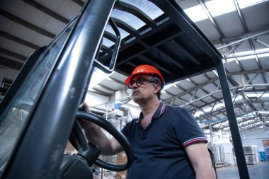 Koruyucu miğferi ve siyah gözlüklü bir işçi depoda forklift kullanıyor.