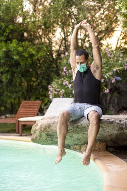 Yazlık kıyafetli, şapkalı ve cerrahi maskeli bir adam otelin havuzunda dinleniyor.