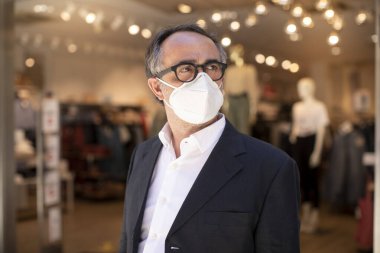 Ceketli ve cerrahi maskeli bir adam şehir merkezindeki bir giyim mağazasının önünde duruyor.