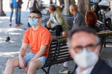 Koruyucu maskeli genç adam şehirde bir bankta oturuyor.