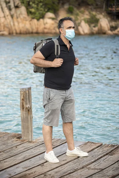 夏の服装で暗い髪の男とバックパックは保護顔マスクと海辺のリゾートタウンを歩いてください — ストック写真
