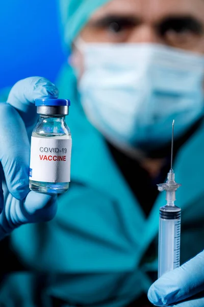 带着外科口罩 护目镜和乳胶手套的男人手里拿着一瓶蓝色背景的抗弧菌疫苗 — 图库照片