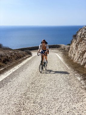 Bisikletli bir kız deniz boyunca giden bir yolun yamacında pedal çeviriyor.