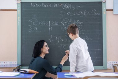 Öğretmen bir çocuğun okul tahtasına matematiksel işlemler yapmasına yardım ediyor.