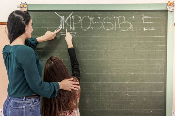 一位穿着绿色衬衫的黑头发老师和她的一个学生一起在黑板上标出了一个写着 不可能 删除了前两个字母 表示一切皆有可能 — 图库照片