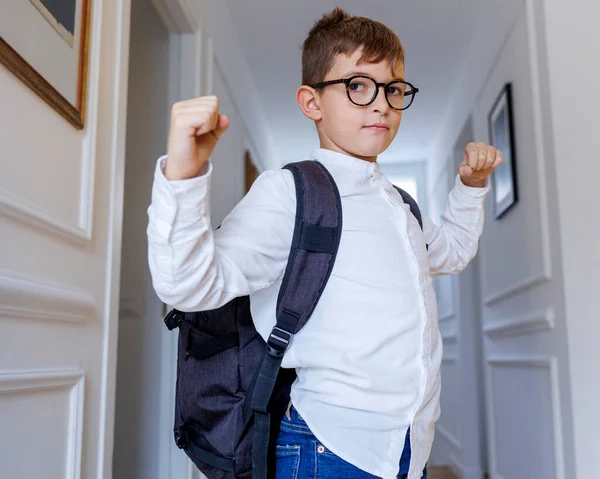 金发碧眼的孩子戴着眼镜 背着背包 站在走廊里 一边玩耍一边表现出肌肉 — 图库照片