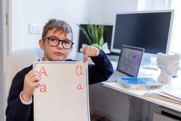 金发男孩戴着眼镜 身穿蓝色衣服 坐在桌子前 他看到一块标有字母字母的板子 桌上有计算机和监视器 — 图库照片